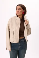 Campera corta color beige claro, estilo bomber o trucker, confeccionada en símil cuero suave. Presenta cuello mao, cierre frontal metálico, bolsillos frontales con tapa y botones a presión, y reguladores ajustables en el ruedo.