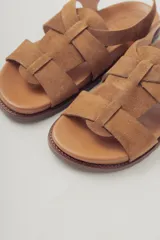 Sandalia plana de gamuza color marrón oscuro, con tiras cruzadas en la parte delantera y una tira fina ajustable con hebilla metálica en el tobillo. Posee plantilla de cuero y suela baja.