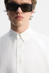 Camisa blanca de corte regular fit confeccionada en popelín de algodón. Tiene cuello abotonado, manga larga con puños abotonados y cierre frontal con botones.