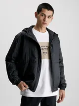 Campera cortavientos negra con capucha, confeccionada en poliéster reciclado. Cuenta con cierre de cremallera, puños y bajo elásticos, bolsillos laterales y logo de Calvin Klein en la manga.