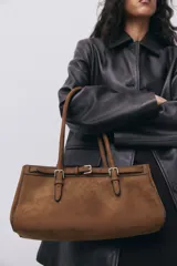 Cartera marrón de gamuza sintética con doble asa corta, cierre magnético y correa decorativa con hebilla metálica.