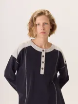 Buzo de manga larga con diseño de bloques de color y textura. El cuerpo principal es marrón, la espalda superior y hombros tienen un panel de tejido a rayas grises y azules, y los hombros superiores son de color beige rosado. Presenta costuras decorativas en rojo que separan los bloques de color.