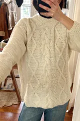 Sweater tejido a mano en lana merino color crudo, con cuello redondo y diseño de trenzas en el frente.