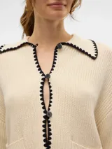 Cardigan tejido color crema con cuello polo, cierre con botones negros y ribete negro con forma de ondas en cuello, cierre, bolsillos y ruedo.