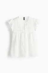 Top blanco de manga corta con escote redondo y abertura en V con tiras para atar. Presenta canesú con inserciones de encaje y costura fruncida con volados. Corte holgado.