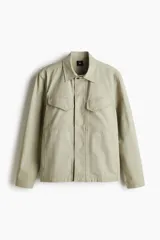 Sobrecamisa color arena de lona gruesa de algodón, corte recto, con cuello clásico, cierre frontal con botones a presión ocultos, canesú en la espalda, dos bolsillos superiores con solapa y botón a presión, bolsillos laterales discretos, mangas largas con puños abotonados y forro interno.