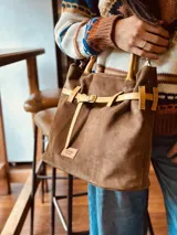 Bolso marrón de cuero gamuza con detalles de correa en cuero mostaza. Tiene una correa corta para llevar como bolso de mano y una correa larga extraíble y regulable. En el interior tiene bolsillo y llavero.