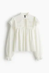 Blusa blanca de manga larga con cuello alto y lazo para atar. Presenta detalles de volados y calados tipo encaje en el canesú y puños con elástico y volado.