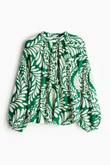 Blusa de tejido liviano color verde con estampado de hojas blancas, cuello corto fruncido y tapeta de botones. Mangas globo largas con volumen de corte raglán y puños finos abotonados.
