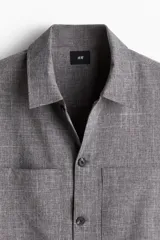Sobrecamisa gris de tejido suave con cuello inglés, tapeta lisa, corte estándar, bolsillos superiores abiertos, abertura con botón en los puños, canesú en la espalda y bajo recto.