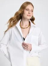 Camisa blanca de corte recto y estilo oversized, con cuello clásico, mangas largas con puños anchos, bolsillo delantero, cierre frontal con botones ocultos y bajo ligeramente redondeado.