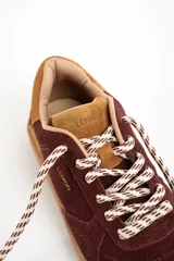 Zapatilla de cuero con capellada de gamuza en tonos borgoña y camel, con suela de goma color caramelo. Cordones trenzados en blanco y marrón.