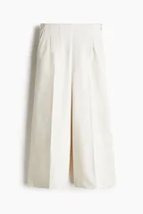 Pantalón palazzo color blanco hueso de tiro alto con elástico oculto en la cintura, cierre lateral oculto y pinzas que realzan la figura. Modelo de corte holgado con bolsillos al bies, bolsillos decorativos atrás y piernas tres cuartos con raya adelante y atrás.
