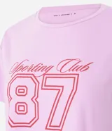 Remera corta de manga corta color rosa claro, con cuello redondo y estampado frontal que incluye el texto "Sporting Club" y el número "87" en rojo.