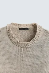 Jersey de punto color beige, con cuello redondo, manga larga y corte holgado. Presenta un lavado con efecto desgastado y rotos en toda la prenda.
