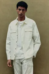 Sobrecamisa color crema con cuello inglés, cierre frontal con botones a presión, dos bolsillos superiores con solapa y puños ajustables con botón a presión.