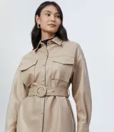 Campera de poliuretano (PU) color beige, con cuello camisa, cierre frontal de botones y dos bolsillos con solapa en el pecho. Incluye un cinturón ajustable con hebilla redonda forrada al tono.