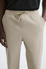 Pantalón de jogging color beige confeccionado en tejido de mezcla de algodón. Cuenta con cintura elástica ajustable con cordón, bolsillos frontales y traseros.