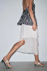 Falda midi color blanco, semitransparente, de tiro medio con cintura elástica, forro corto interior y abertura en la espalda.
