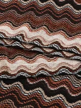 Bufanda tipo chalina de tejido de punto con diseño de ondas en tonos marrón, beige y negro. Tiene flecos en los extremos.
