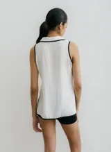 Blusa color beige con cuello camisero y botones frontales, sin mangas y con ribete negro en cuello, sisas y ruedo.