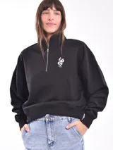 Buzo negro de corte cropped con cuello alto y cierre metálico plateado hasta el pecho. Tiene un pequeño bordado blanco con forma de serpiente en el lado izquierdo del pecho.