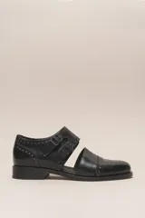 Zapato bajo de cuero negro, estilo monk strap con dos hebillas ajustables sobre el empeine. Presenta detalles de brogue (perforaciones) en la puntera y alrededor del borde superior. Tiene un taco bajo y ancho.
