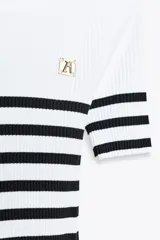Camiseta de rib a rayas horizontales blancas y negras, de corte ajustado, cuello redondo y manga corta. Presenta un broche metálico en el frente y una medalla con el logo de Zara en la espalda.