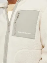Campera de teddy color blanco hueso, de manga larga y cuello alto. Tiene cierre con cremallera, bolsillos frontales y un bolsillo gris en el pecho con cierre.