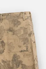 Pantalón de jean color beige con estampado abstracto floral en tonos marrones, de corte amplio, con bolsillos delanteros y traseros, y detalle de tira multifunción en la pierna. Cierre frontal con cremallera y botón.