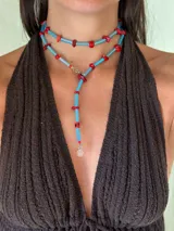 Collar largo estilo lariat o yoyó, compuesto por bastones de vidrio facetados color celeste/azul claro y cuentas de piedra coral/rojo. Cuenta con herrajes de acero quirúrgico y un dije colgante con un logo grabado.