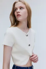 Saco corto color blanco hueso, de punto elástico con textura. Tiene cuello en V, mangas cortas, cierre frontal con tres botones grandes y abertura en el bajo.