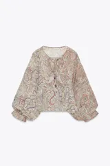 Camisa estampada con motivos paisley en tonos beige, rosa, lila y amarillo. Tiene cuello redondo con lazos, mangas largas abullonadas con puños elásticos y volantes en el delantero.