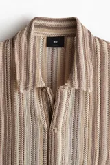 Camisa de manga corta con cuello inglés, botones adelante y bajo recto. Tejido de punto pointelle con estilo crochet y diseño a rayas verticales en tonos beige, marrón y blanco.