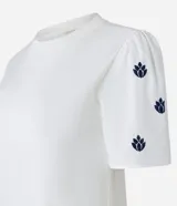 Blusa blanca de algodón con cuello redondo y mangas cortas abullonadas. Presenta pequeños bordados en color azul marino distribuidos en las mangas.