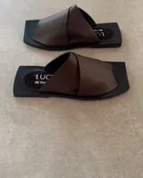 Sandalias chatas de cuero vacuno color marrón con base negra. Diseño con tira ancha cruzada sobre el empeine. Producto de industria uruguaya.