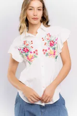 Camisa blanca de manga corta con bordado de flores en el frente. Tiene cuello clásico y cierre frontal con botones. Las mangas son cortas con volados.
