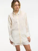 Camisa de manga larga color crema, con cuello camisero y botones frontales. Presenta apliques bordados en la zona del pecho y hombros. El ruedo es irregular, más largo en la parte trasera.