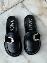 Zuecos negros de cuero con suela gruesa, detalle de tira de gamuza con hebilla metálica plateada y logo "AMÉN" estampado en la plantilla.