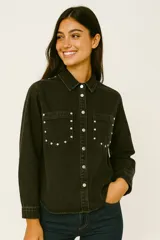 Camisa de jean negra con cuello clásico, cierre frontal con botones a presión, dos bolsillos con solapa en el pecho y tachas plateadas decorativas en cuello, hombros y bolsillos.