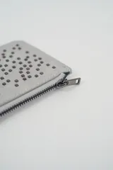 Billetera pequeña color gris claro con cierre de cremallera perimetral y tachas metálicas oscuras distribuidas en el frente.