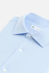 Camisa celeste a rayas verticales blancas, de corte recto, con cuello italiano abierto, manga larga con puños abotonados y cierre frontal con botones.