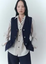 Chaleco de denim azul oscuro con cuello en pico, sin mangas, dos bolsillos delanteros y cierre con botones.