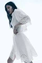 Vestido corto blanco de crepé con secciones de broderie, cuello camisero, botones ocultos, mangas cortas abullonadas, cinturón extraíble y ribete festoneado.