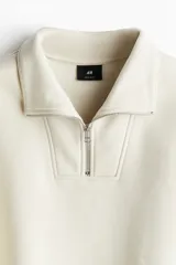 Buzo color crema de tejido pesado con interior cepillado, cuello elevado, medio cierre y hombros caídos. Puños y bajo en punto acanalado. Corte estándar.