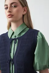 Chaleco tejido de punto color beige con trama texturada y diseño liviano. Presenta cuello a la base, silueta recta y cierre frontal mediante tres lazos delicados.