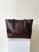 Cartera tipo tote color beige, hecha en cuero graneado con textura uniforme y suave al tacto. Tiene doble asa de hombro, un bolsillo frontal con broche imantado, un bolsillo trasero con cierre metálico y cierre principal también metálico. El interior está forrado y cuenta con tres bolsillos: uno con cierre, uno tipo plaqué para celular y un bolsillo grande tipo sobre con solapa y botón.