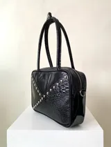 Cartera rígida negra de cuero con tachas plateadas y tres combinaciones de cuero diferentes que aportan textura. Tiene herrajes y cierre metálico plateado marca YKK. Cuenta con un asa fina para llevar al hombro o en la mano y una correa larga extra regulable.