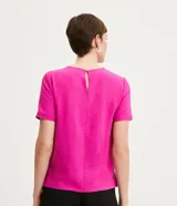 Blusa blanca de poliéster con cuello redondo, mangas cortas, cierre trasero en forma de gota, pequeñas aberturas laterales y acabados en pespuntes en las mangas y el cuello.