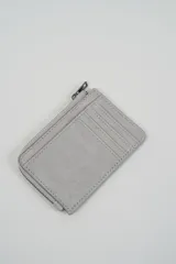 Billetera pequeña color gris claro con cierre de cremallera perimetral y tachas metálicas oscuras distribuidas en el frente.